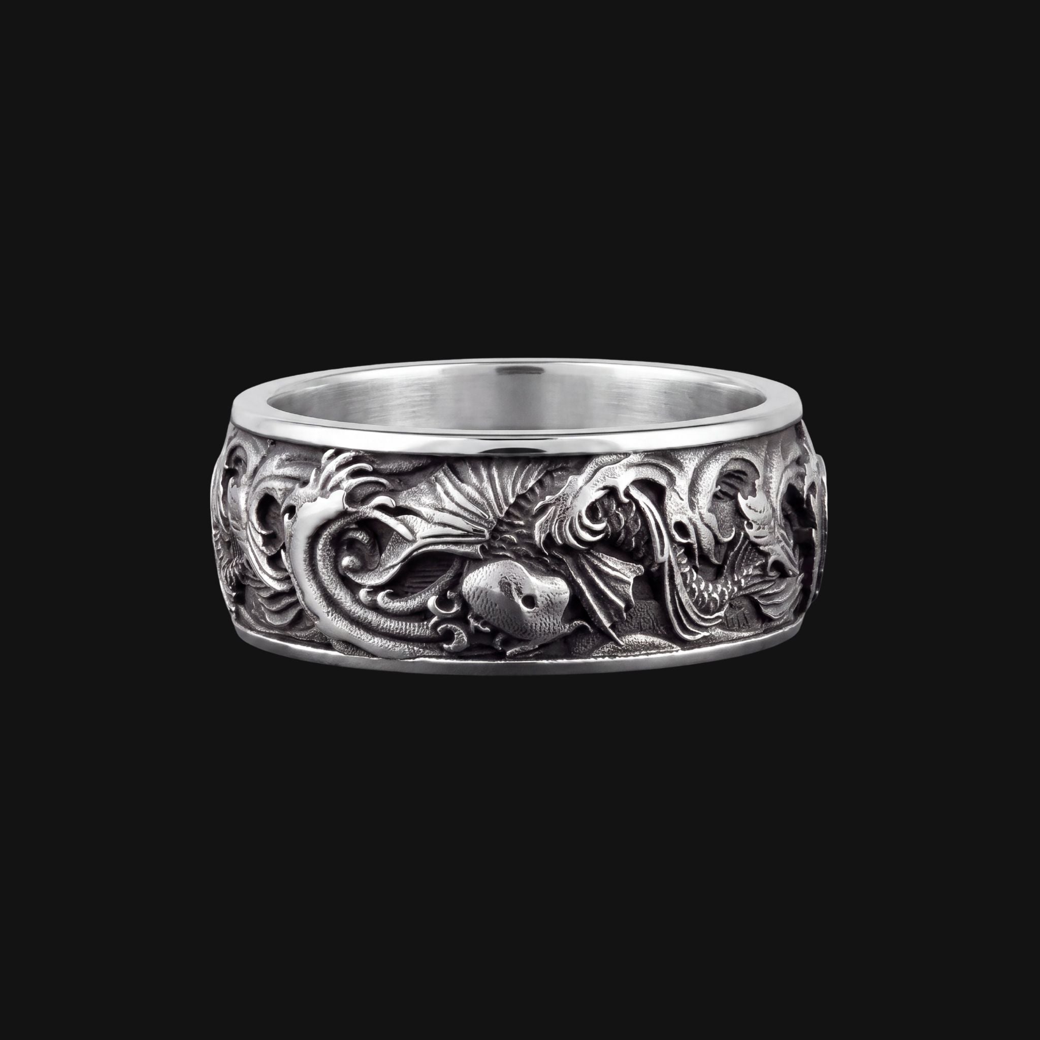 A wide sterling silver band ring featuring intricate 3D relief carvings of dragons and sea creatures, shown against a solid black background.