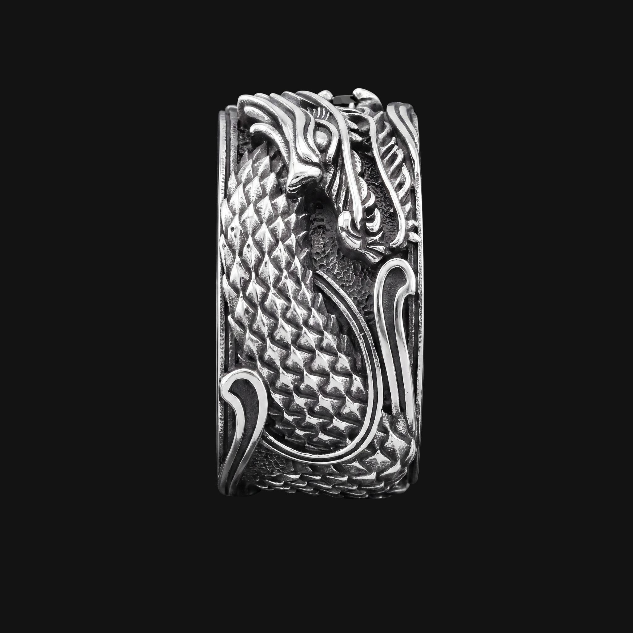 A silver-toned metal ring featuring a highly detailed, raised dragon design with textured scales and flowing decorative lines, shown against a solid black background.